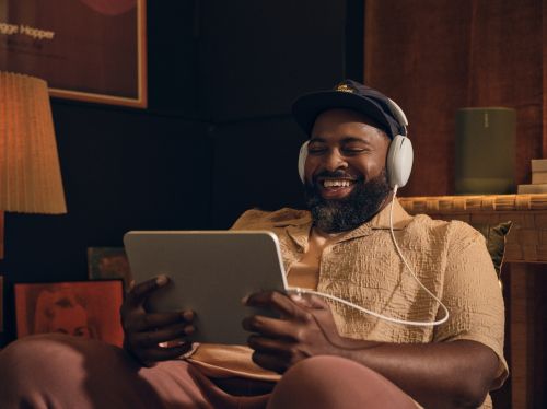 Bearded adult male smiles and wears headphones while looking at a tablet indoors, dressed casually with warm lighting and a poster in view.