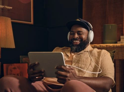 Bearded adult male smiles and wears headphones while looking at a tablet indoors, dressed casually with warm lighting and a poster in view.