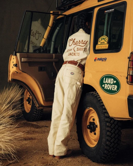 A person in a white shirt and jeans standing beside a yellow Land Rover on a dirt surface in an outdoor environment with tall grass.