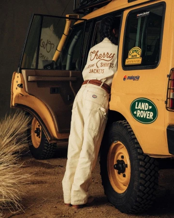A person in a white shirt and jeans standing beside a yellow Land Rover on a dirt surface in an outdoor environment with tall grass.