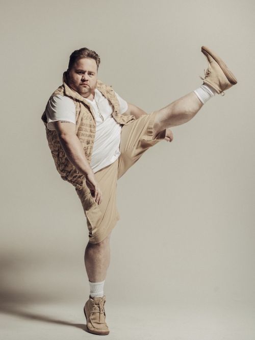 Man in beige shorts, white t-shirt, and vest raises a leg, showcasing casual fashion against a neutral backdrop.
