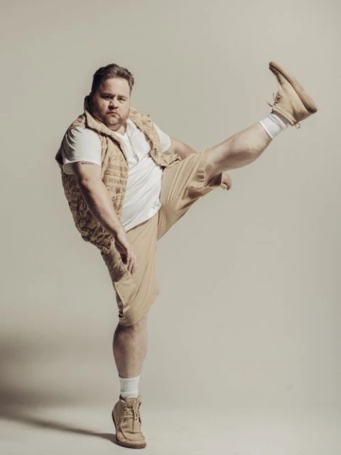 Man in beige shorts, white t-shirt, and vest raises a leg, showcasing casual fashion against a neutral backdrop.