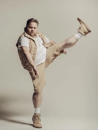 Man in beige shorts, white t-shirt, and vest raises a leg, showcasing casual fashion against a neutral backdrop.