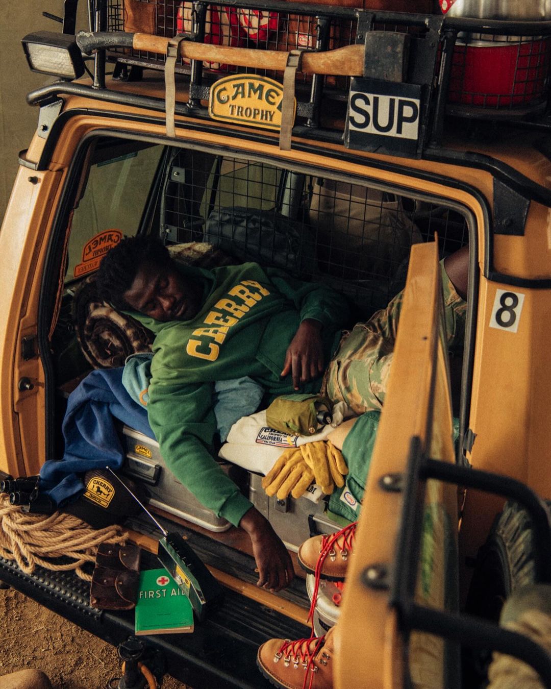 Man asleep inside the back of an off-road vehicle with various camping and outdoor adventure equipment visible in a natural light setting.