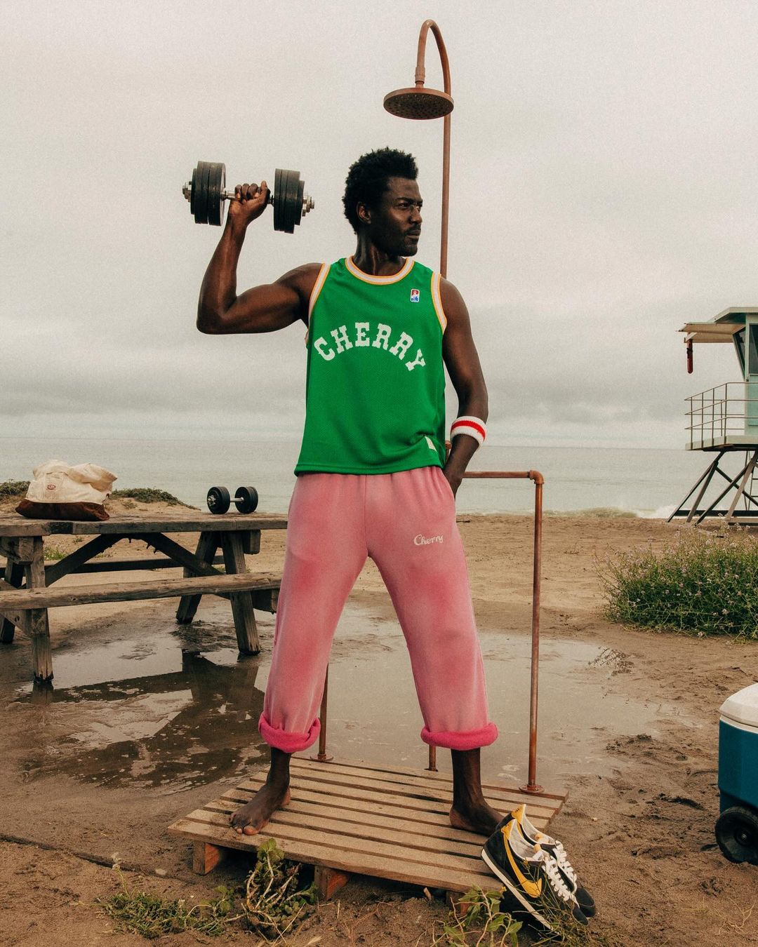African American man lifts dumbbell on a beach next to outdoor shower wearing Cherry brand apparel with ocean and lifeguard tower.