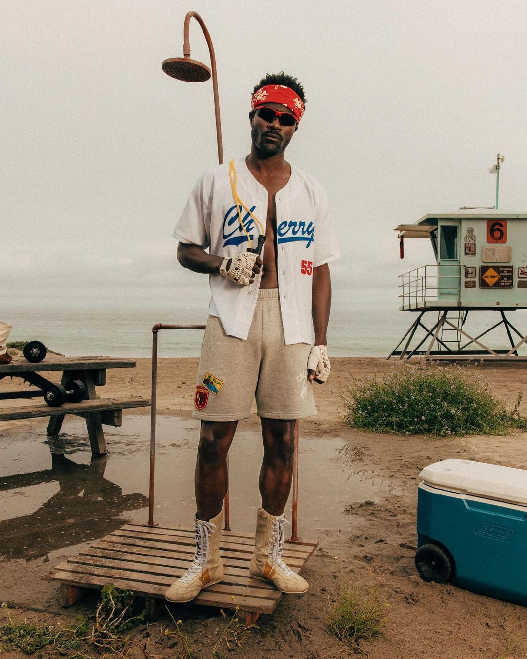 Adult male stands on sandy beach near lifeguard tower, wearing baseball jersey, shorts, and boxing boots, holding jump rope.