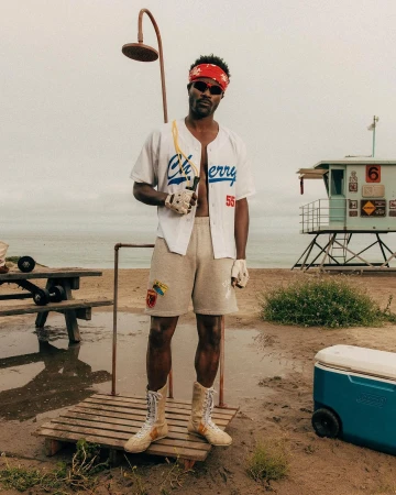 Adult male stands on sandy beach near lifeguard tower, wearing baseball jersey, shorts, and boxing boots, holding jump rope.