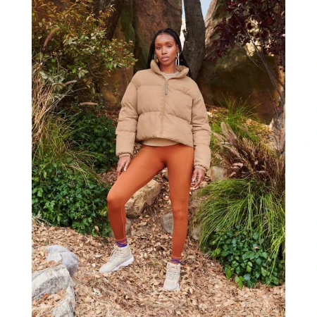 Woman in puffer jacket and leggings poses outdoors amid natural setting with trees and rocks, complemented by muted color palette.