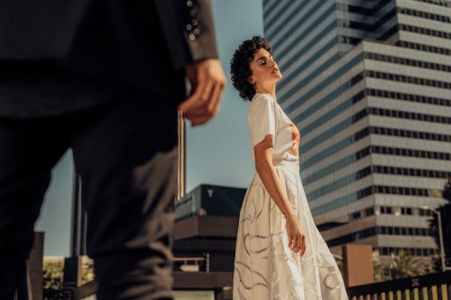 Woman in a white dress gazes up towards sunlight with a city building backdrop. A man in a dark suit is in the foreground.