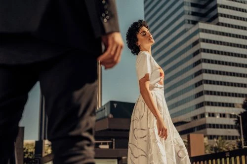 Woman in a white dress gazes up towards sunlight with a city building backdrop. A man in a dark suit is in the foreground.