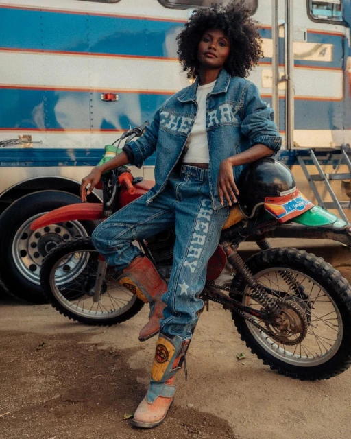 A Black woman poses wearing a Cherry-branded denim outfit, leaning against a red motorcycle with a blue and white trailer in the background.