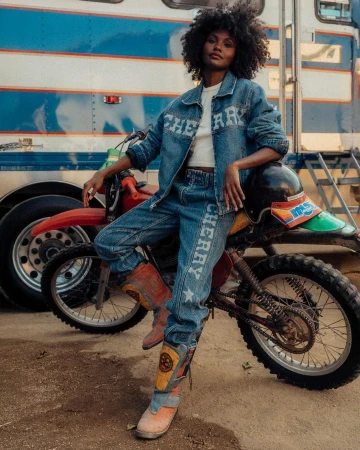 A Black woman poses wearing a Cherry-branded denim outfit, leaning against a red motorcycle with a blue and white trailer in the background.