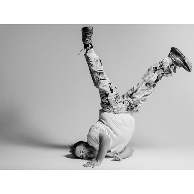 Monochrome studio shot of a male adult in a handstand wearing patterned pants and sneakers against a gray background.