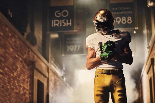 Football player in uniform with a helmet, gold pants, and green gloves stands in a brick tunnel with signage in the background.