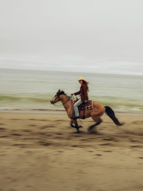 A woman in a cowboy hat rides a tan horse at a gallop on a sandy beach with waves in the background under an overcast sky.