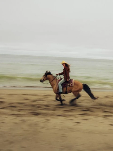 A woman in a cowboy hat rides a tan horse at a gallop on a sandy beach with waves in the background under an overcast sky.