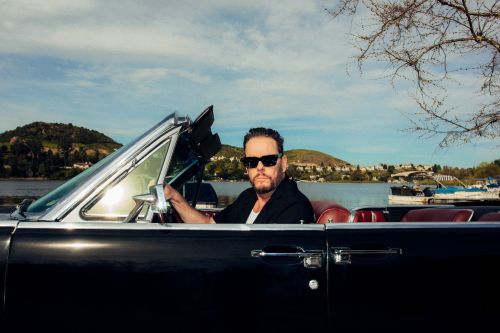 Man wearing sunglasses seated in a classic convertible black car by a lake under an open sky