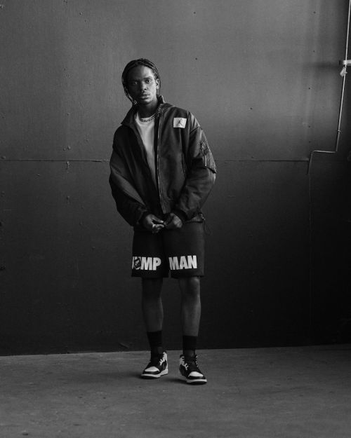 Black and white portrait of a young adult male in a jacket and shorts posing against a wall inside a photo studio.