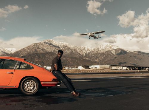 A man leans against an orange Porsche on an asphalt runway with a small airplane taking off into a partly cloudy sky with mountains.
