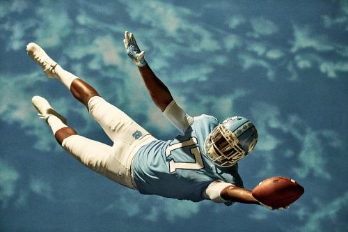 American football player in blue uniform leaps forward, reaching out to catch a brown football against a blue sky backdrop with subtle clouds.