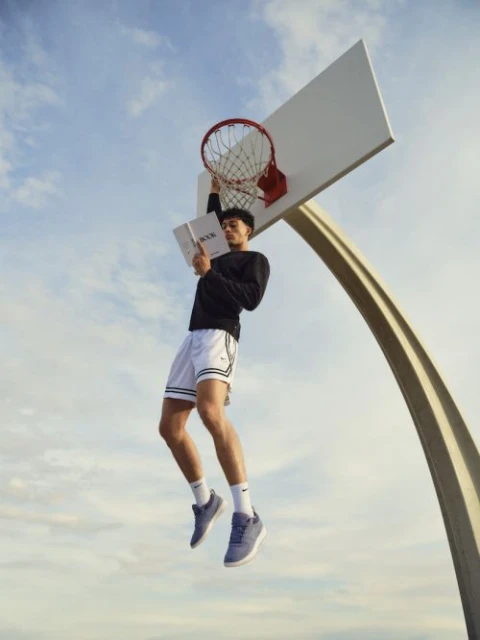A man in sportswear hangs from a basketball hoop while reading a book, set against a bright blue sky with soft, wispy clouds.