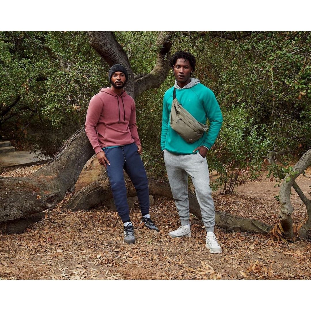 Two adult men pose outdoors in hooded sweatshirts and athletic pants amid greenery and fallen leaves, with a modern casual style.