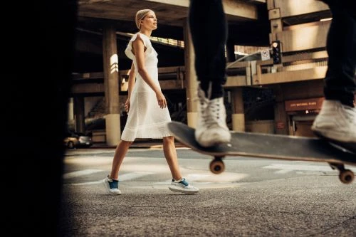Woman in a white dress crosses a city street as a person on a skateboard rides past in blurred motion on an urban crosswalk.