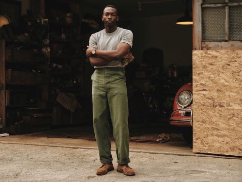 Man in gray tee and green pants stands in garage with red car, arms crossed, looking at the camera in a medium, full shot.