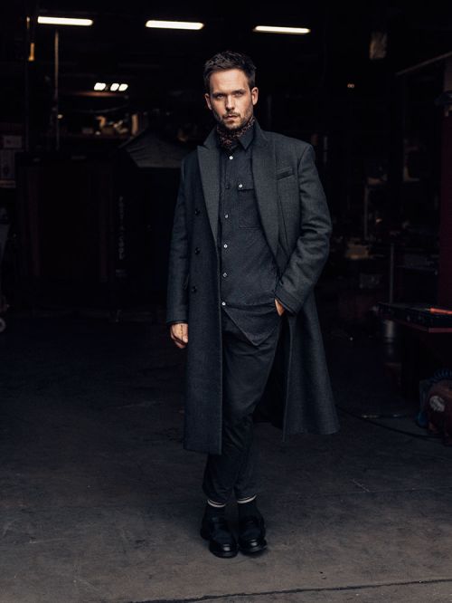 Full-length portrait of a man in a dark coat and scarf, standing in an industrial setting, captured with natural lighting.