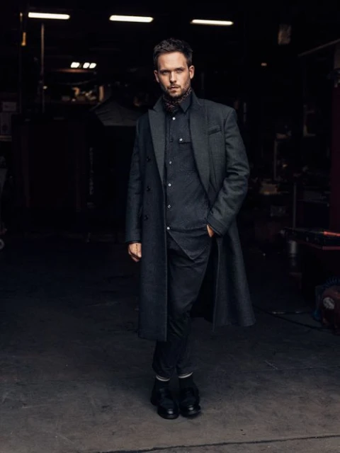 Full-length portrait of a man in a dark coat and scarf, standing in an industrial setting, captured with natural lighting.
