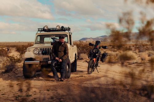 An adult man sits on the hood of a vintage pickup truck parked near a black and orange motorcycle in a desert landscape under a partly cloudy sky.