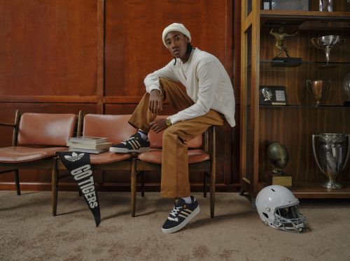 An adult male wearing casual attire poses in a retro-style room with athletic memorabilia and adidas sneakers while sitting on a chair.