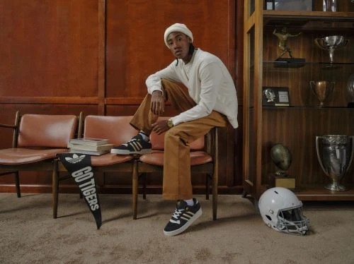 An adult male wearing casual attire poses in a retro-style room with athletic memorabilia and adidas sneakers while sitting on a chair.