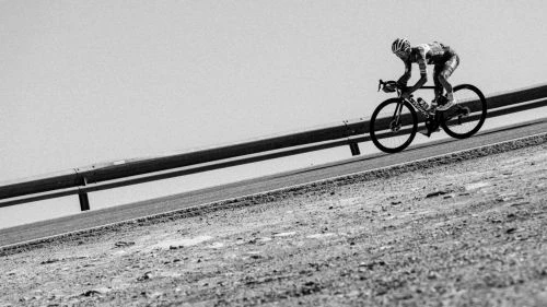 Two cyclists in matching outfits ride bicycles on a paved road next to a guardrail, captured in black and white under a bright sky.