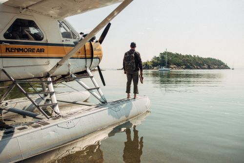 A man standing on a floatplane's pontoon, looking out at a forested island and sailing boat in the distance on a sunny day.