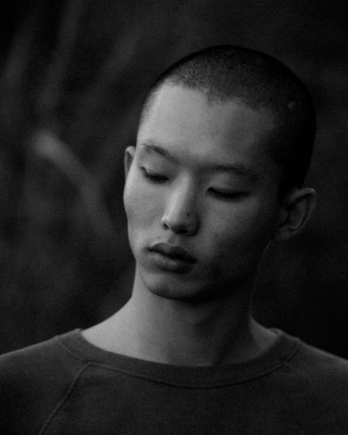 Portrait of a young adult with a shaved head, eyes closed, and wearing a dark sweater in a dark, natural setting; black and white.