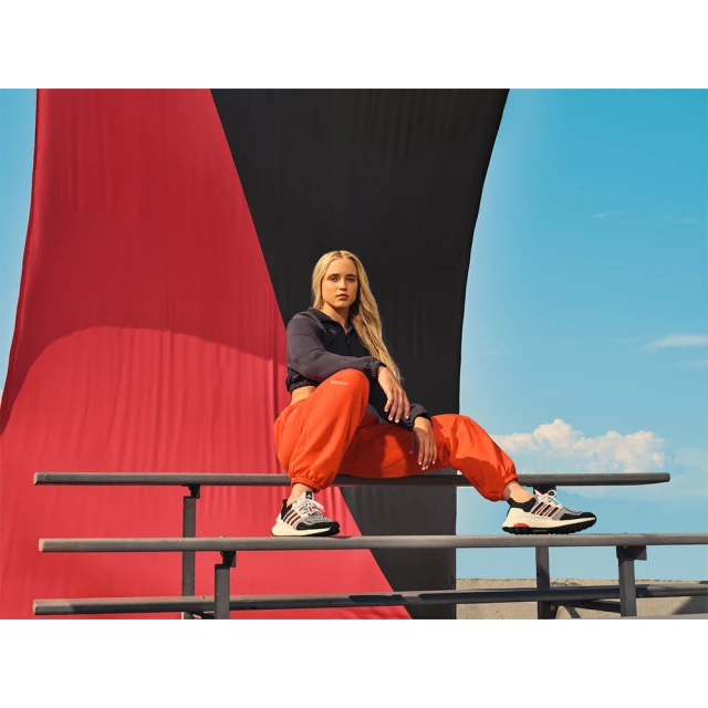 Athletic woman with long blonde hair sits in orange sweatpants and running shoes on railing against backdrop of sky and black/red panels.