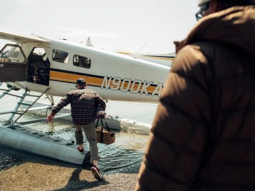 Adult male boarding a small aircraft on the water with a duffel bag. Another man is walking toward the airplane, near the shore.
