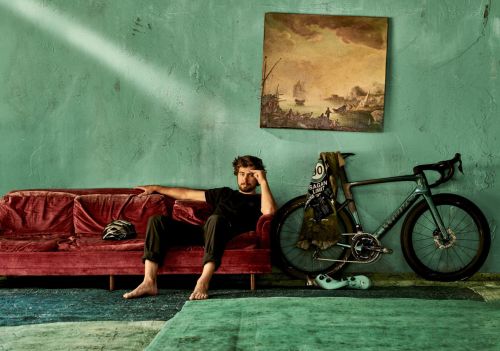 Adult male seated on a velvet couch next to bicycle. Artwork and wall are in the background in an indoor setting.