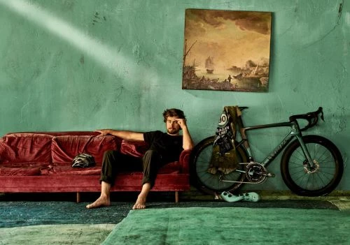 Adult male seated on a velvet couch next to bicycle. Artwork and wall are in the background in an indoor setting.