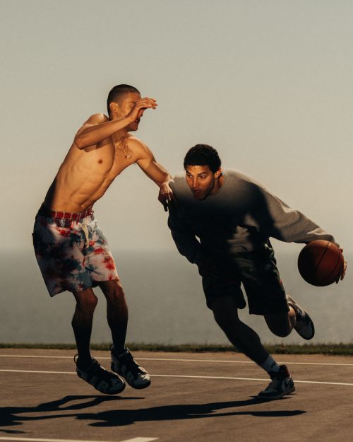 Two men playing basketball outdoors under bright sky, one shirtless in patterned shorts, the other in a sweatshirt, action shot.