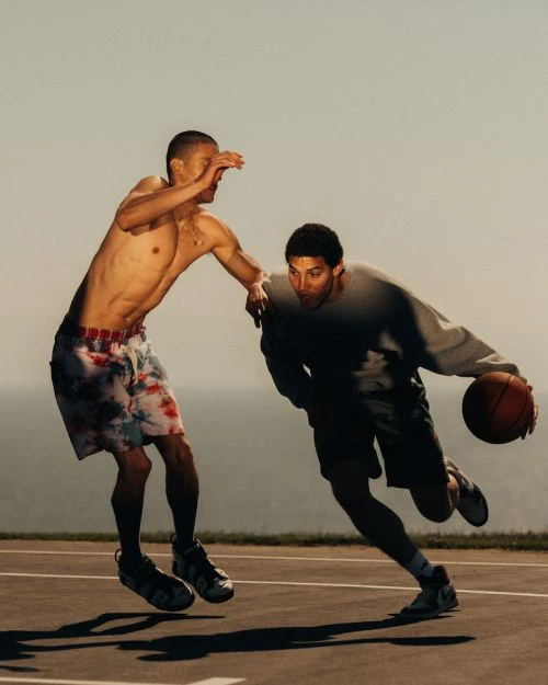 Two men playing basketball outdoors under bright sky, one shirtless in patterned shorts, the other in a sweatshirt, action shot.