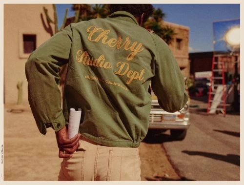 A man in an olive jacket with gold lettering stands on a sunny street with a vintage car, buildings, and film equipment in the background.