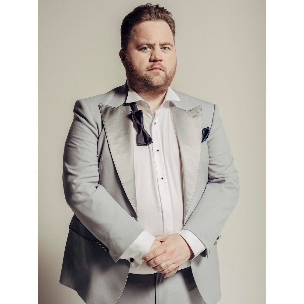 Portrait of an adult male in a light grey suit and loosened bow tie against a neutral backdrop, arms crossed, looking at the camera.