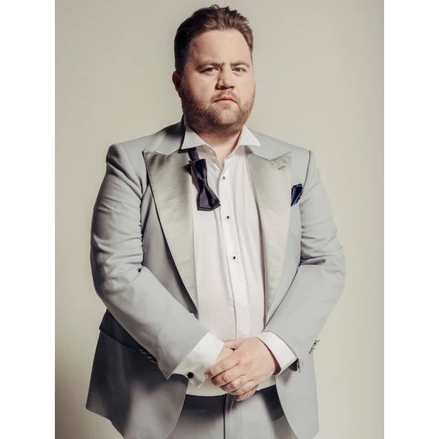 Portrait of an adult male in a light grey suit and loosened bow tie against a neutral backdrop, arms crossed, looking at the camera.