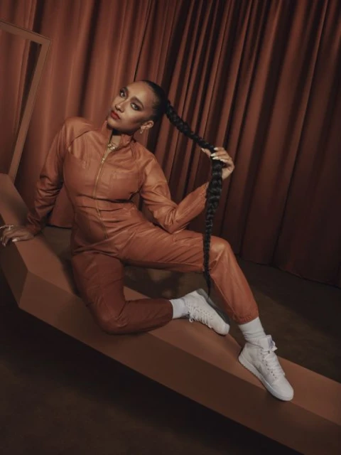 A woman with a long braid poses in a brown jumpsuit and white sneakers against a brown backdrop, seated on a brown geometric shape.