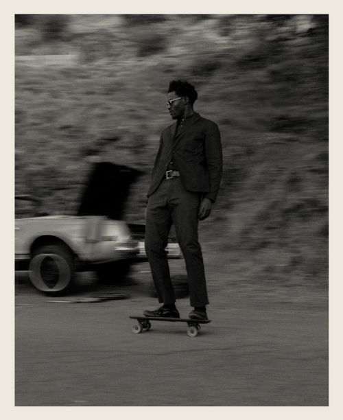 Black man riding a skateboard wearing a dark suit jacket and trousers with a parked car in the blurred background