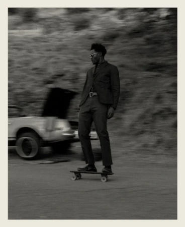 Black man riding a skateboard wearing a dark suit jacket and trousers with a parked car in the blurred background