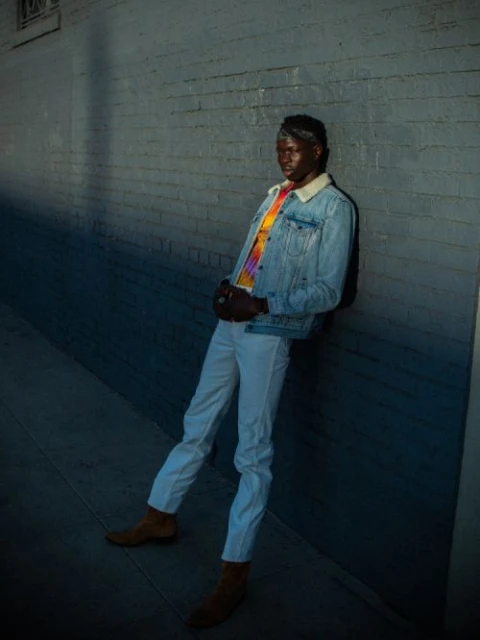 A Black male model in a denim jacket and white pants leans against a wall, casting a shadow, and creating a fashion-forward urban portrait.