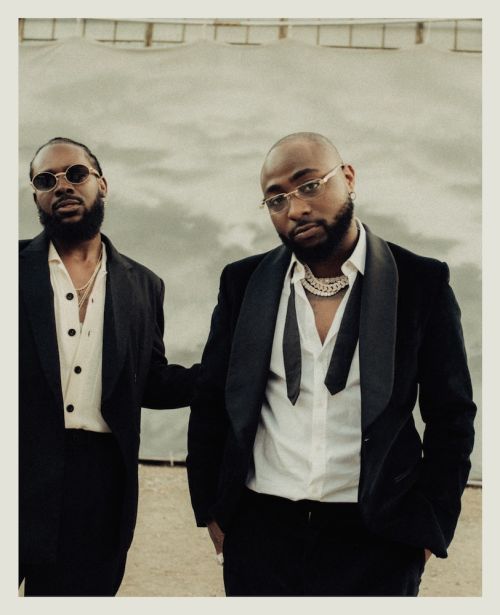 Two adult males pose in formal black suits against a textured cloudy background. One man wears round glasses; the other a chain necklace.